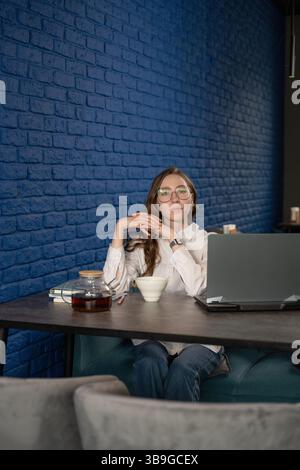 Eine Frau mit Brille sitzt an einem Café-Tisch mit Laptop, Teetasse und Teekanne vor einer stilvollen blauen Ziegelwand und macht eine entspannende Pause von der Arbeit. Stockfoto