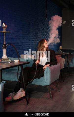 Eine Frau sitzt auf einem Sessel in einem schwach beleuchteten Raum und atmet anmutig Rauch aus einer Wasserpfeife aus. Die Umgebung verströmt Ruhe mit seinem gemütlichen Ambiente und Stockfoto