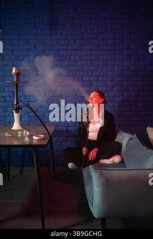 Eine Frau sitzt auf einem Sofa und genießt ruhig eine Wasserpfeife mit Rauch in einer gemütlichen Lounge. Sie ist umgeben von Kissen und einem blauen Mauerrücken Stockfoto