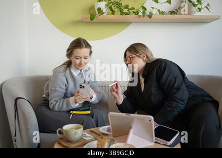 Zwei Frauen, die sich bei Kaffee in einem modernen Café unterhalten. Sie diskutieren über die Arbeit, mit einem Smartphone und Tablet auf dem Tisch, Surround Stockfoto
