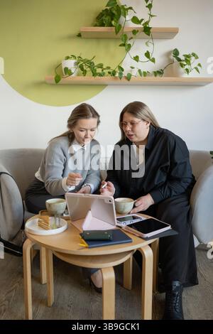 Zwei Frauen nehmen an einer gemeinsamen Diskussion in einem gemütlichen Café Teil, indem sie ein Tablet verwenden. Sie sitzen an einem Holztisch mit Kaffeetassen, umgeben von Grün und Stockfoto