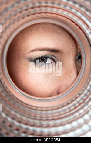 Eine Nahaufnahme eines weiblichen Auges, der durch die kreisförmige Öffnung einer strukturierten Pfeife schaut und die Details ihres Make-ups, der Hautstruktur und der Pfeife festnimmt Stockfoto