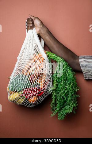 Eine Person hält einen wiederverwendbaren Netzbeutel mit frischem Gemüse, einschließlich Karotten und Paprika, vor braunem Hintergrund. Fördert nachhaltiges Geschäft Stockfoto