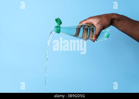 Nahaufnahme einer Hand, die Wasser aus einer durchsichtigen Plastikflasche vor hellblauem Hintergrund gießt. Das Bild erfasst die fließende Bewegung und Transparenz o Stockfoto