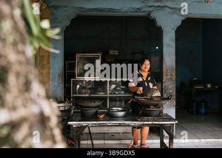 Eine Frau in einem Blumenkleid kocht köstliche traditionelle Gerichte in einem Marktstand im Freien. Die Szene fängt das Wesen der Street Food-Kultur und des Culin ein Stockfoto