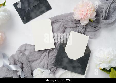 Elegantes Hochzeitsgeschirr mit dunklen Umschlägen und leeren Karten. Begleitet von weichem grauem Stoff und weißen Pfingstrosenblüten, schaffen Sie eine raffinierte Stockfoto