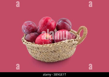 Reife rote Pflaumen, die aus einem kleinen Korb überlaufen, schaffen ein farbenfrohes Stillleben vor einem leuchtend rosa Hintergrund Stockfoto