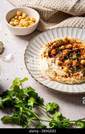 Ein Teller mit cremigem Hummus, garniert mit Kichererbsen, Kräutern und Gewürzen steht auf einem strukturierten Hintergrund. Eine Schüssel mit Kichererbsen und frischen Kräutern bereichert das VI Stockfoto