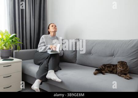 Eine Frau sitzt auf einem grauen Sofa, hält ein Buch, sieht friedlich und besinnlich aus. Neben ihr liegt eine Katze ruhig. Eine grüne Pflanze und eine Schublade werden zur hinzugefügt Stockfoto