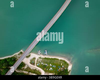 Bild der Drohne von oben nach unten vom Castreccioni-See mit einer Brücke, die türkisfarbenes Wasser und einen kleinen Strandbereich mit angedockten Booten überquert, was eine ruhige, organisierte Atmosphäre schafft Stockfoto