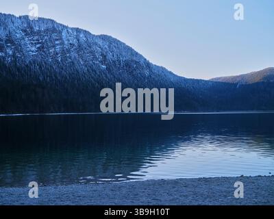 Ruhiger Blick auf einen schneebedeckten Berg, der sich in der Abenddämmerung auf ruhigem Seenwasser in der Nähe eines Kieselstrandes spiegelt Stockfoto