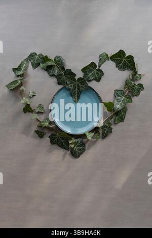 Eine Herzform aus Efeublättern vor grauem Hintergrund und einer blauen Platte in der Mitte, eine nachhaltige symbolische Dekoration zum Thema Liebe Stockfoto