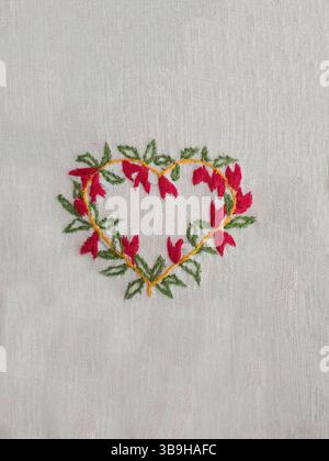 Postkartenmotiv mit gesticktem Herz aus grünen Blättern und roten Blumen auf weißem Stoff Stockfoto