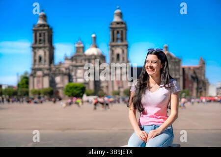 Junge Latina-Frau mit langen Haaren, rosa-weißem T-Shirt und Jeans, die tagsüber im Zocalo von Mexico City sitzt. Stockfoto