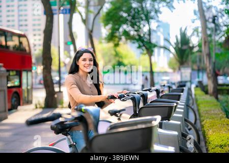 Die junge Latina in lässiger Kleidung lächelt, während sie eine öffentliche Fahrradstation in Ecobici an der Reforma Avenue in Mexiko-Stadt benutzt. Stockfoto