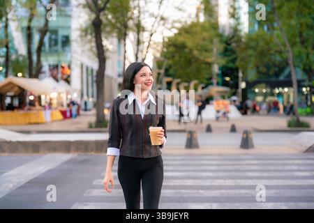 Die junge Latina lächelt selbstbewusst, während sie über die Avenida Reforma in Mexiko-Stadt läuft und bei Abendlicht einen Eiskaffee hält. Stockfoto