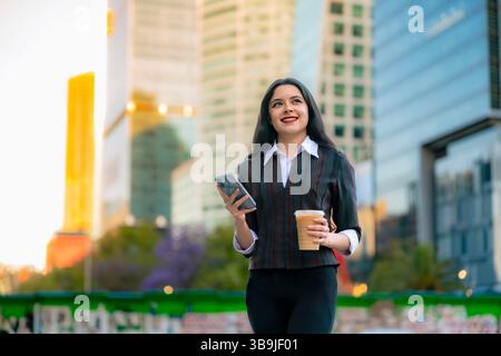 Latina in formellem legeren Outfit sitzt draußen auf Reforma, lächelt, während sie ihr Telefon überprüft und in Mexiko-Stadt Eiskaffee hält. Stockfoto