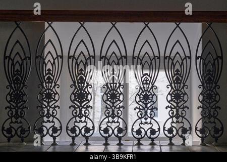 Dekorative schmiedeeiserne Geländer mit komplizierten Blumen- und geometrischen Motiven, die vor einem hellen Fenster in traditioneller Inneneinrichtung gehalten werden. Stockfoto