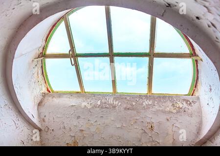 Verwittertes kreisförmiges Fenster mit abblätternder weißer Farbe und grünem Rahmen, das durch die leicht schmutzigen Scheiben einen bewölkten Himmel freigibt. Fenster veraltet, strukturiert Stockfoto
