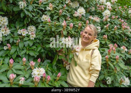 Eine Frau in einem gelben Hoodie lächelt, während sie neben einem üppigen Rhododendronstrauch in voller Blüte steht, umgeben von leuchtenden rosa und weißen Blumen. Sie Stockfoto