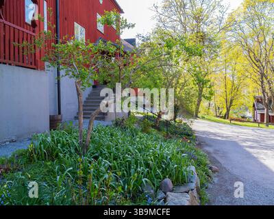 Ein malerischer Blick auf ein bezauberndes rotes Haus mit einer Treppe, die hinauf führt, umgeben von üppigem Grün und blühenden Blumen. Das Sonnenlicht filtert durch den Stockfoto