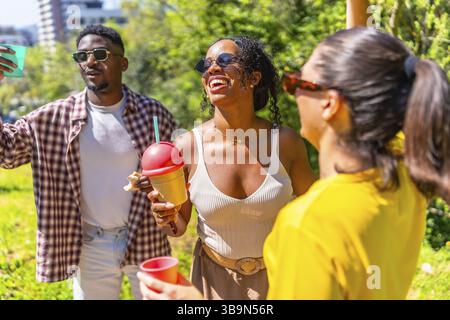 Fröhliche Gruppe von Freunden, die lachen, tanzen und erfrischende Getränke in einer lebhaften Parklandschaft genießen Stockfoto