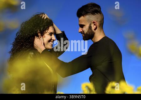 Porträt eines jungen lächelnden Paares, bei dem der Haarmann das Gesicht seiner Freundin streichelt, bevor er blauen Hintergrund und gelbe Blumen küsst Stockfoto
