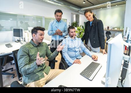 Verschiedene Geschäftsteams arbeiten an einem Computerprojekt zusammen, demonstrieren Inklusion und Teamarbeit am Arbeitsplatz mit einem behinderten Kollegen im Laufrad Stockfoto