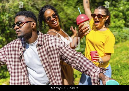 Drei Freunde genießen die Sommersonne, tanzen fröhlich, trinken kalte Getränke und lachen in einer lebhaften Umgebung im Freien Stockfoto
