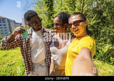 Drei fröhliche, ethnische Freunde machen ein Selfie in einem Park und genießen einen sonnigen Tag zusammen Stockfoto