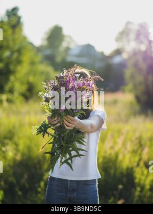 Junge Frau hält wilde Blumenstrauß im Freien in wunderschönem Hintergrundlicht Sonnenuntergang, in der Nähe von grünem Feld auf dem Land. Sommer, ländliches Leben und Naturheilkunde Stockfoto