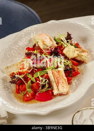 Köstlicher Krabbensalat mit Meeresfrüchten auf weißem Teller auf dem Hintergrund des Restauranttisches. Teller mit gesundem Salat aus Krabben, Tomaten und frischem Gemüse, Dekora Stockfoto