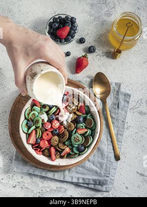 Trendiges Essen - Pfannkuchen-Müsli. Milchgießen auf einen Haufen mehrfarbiger Mini-Müslipfannkuchen in einer Schüssel. Winzige Müslipfannkuchen mit Beeren auf Bastelplatte über g Stockfoto
