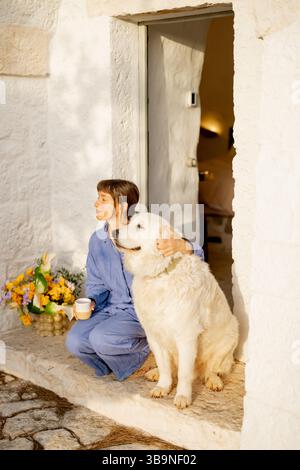 Ein ruhiger Moment, der zwischen einer Frau in blauem Leinen und ihrem treuen weißen Hund geteilt wird, während sie einen friedlichen Sonnenaufgang vor der Haustür eines traditionellen trullo-Hauses in Apulien, Italien, genießt Stockfoto