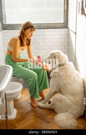 Ein zärtlicher Moment zwischen einer Frau und ihrem großen weißen Hund im Badezimmer, während sie sein Fell sorgfältig mit Liebe und Sorgfalt putzt Stockfoto