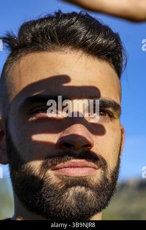 Vertikale Nahaufnahme eines lächelnden Bart-Mannes, der während des Sonnenuntergangs in einem gelben Feld die Sonne mit seiner Hand bedeckt Stockfoto