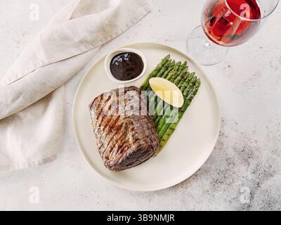 Gegrilltes Fleischsteak auf Teller mit Zitrone und Spargel auf weißem Tischhintergrund. Food and Cuisine Konzept. Verzierung im Restaurant-Stil mit Rotweinglas Stockfoto