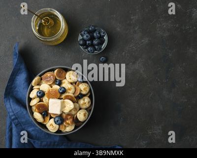 Trendiges Essen: Pfannkuchen, Müsli, Butter und Heidelbeeren. Haufen Mini-Cerealien-Pfannkuchen in Boul auf dunklem Hintergrund. Platz rechts für Text kopieren. Obere V Stockfoto