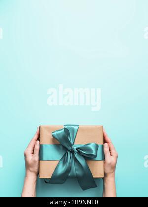 Weibliche Hände halten große Geschenkbox auf türkisblauem Hintergrund, kopieren Raum. Kaukasische Mädchen Hände halten Geschenkbox in Handwerk Verpackung Papier mit grünen sati Stockfoto