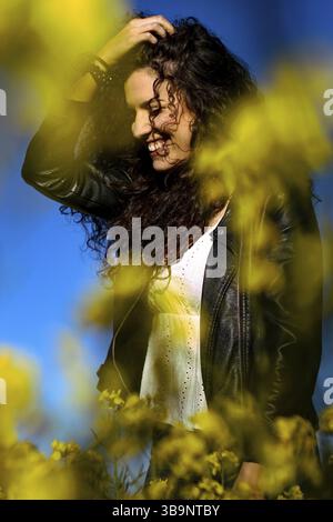 Vertikales Nahporträt einer jungen Frau, die ihre Hand auf dem Kopf hält und an einem sonnigen Frühlingstag in einem gelben Blumenfeld lächelt Stockfoto