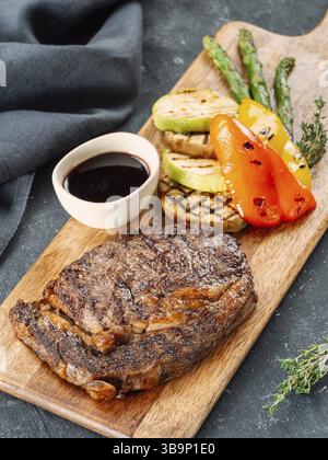 Frisches, heißes Schweinefleisch-Steak auf hölzernem Schneidebrett, serviert Gemüse und Sauce. Köstliches Schweinefleisch auf dunklem Tisch. BBQ Rib Beefsteak oder zartes Schweinefleisch Steak Stockfoto