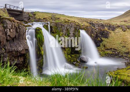 Kirkjufellsfoss Wasserfälle Landschaft, Frühling, Langzeitbelichtung mit Brücke und Felsen bedeckt mit Moos, Snaefellsnes Halbinsel, Island. Stockfoto