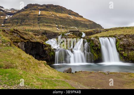 Kirkjufellsfoss Wasserfälle Landschaft, Frühling, Langzeitaussicht mit Brücke und Felsen bedeckt mit Moos und vulkanischen Gebirgszügen, Island. Stockfoto