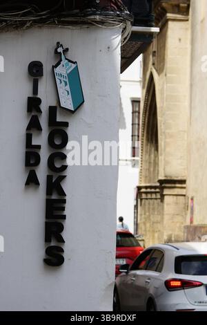Geschäftsschild für Gepäckaufbewahrung mit der Aufschrift „IRALDA SCHLIESSFÄCHER“ an einer weißen Wand in Sevilla, Spanien, mit Autos und historischer Architektur im Hintergrund. Stockfoto
