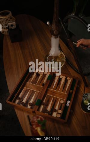 Gemütliche Lounge mit Shisha-Einrichtung und hölzerner Backgammon-Brett, kombiniert mit bunten Cocktails und dunkler Beleuchtung. Stockfoto