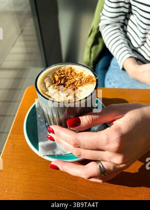 Frau, die im Café Kaffeepause macht, heiße Latte mit Milchschaum trinkt und Sonnenlicht durch das Fenster fängt. Vertikal Stockfoto