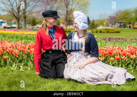 Holland, MI, USA - 24.04.2025: Porträt lächelnder Teenager-Mädchen in traditioneller niederländischer Kleidung, die an sonnigem Tag im blühenden Tulpenfeld stehen und Kultu zeigen Stockfoto