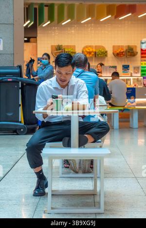 Ein junger asiatischer australischer Mann sitzt mit seinem Telefon und isst in einem Food Court in Sydney, Australien, Mittagessen zum Mitnehmen Stockfoto