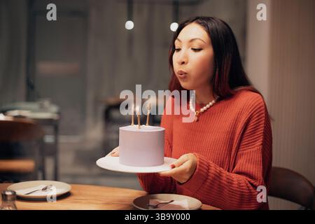 Eine junge Frau feiert ihren Geburtstag und bläst Kerzen auf einem leuchtenden lila Kuchen aus, während sie einen festlichen Abend in einem Restaurant genießt Stockfoto