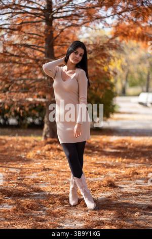 Junge Frau posiert im Herbst in einem Park, trägt ein beiges Kleid und Cowboystiefel, mit gefallenen Blättern auf dem Boden Stockfoto